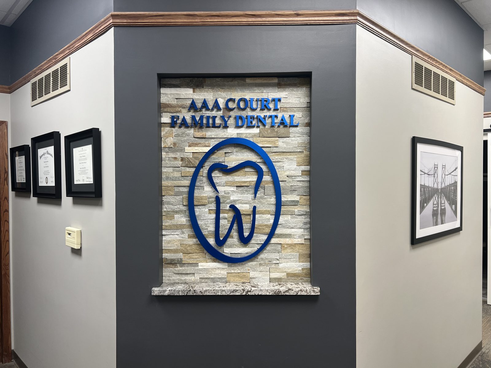 The AAA Court Family Dental interior signature wall, stacked stone, navy raised lettering, the practice's tooth logo, and the doctors' framed diplomas